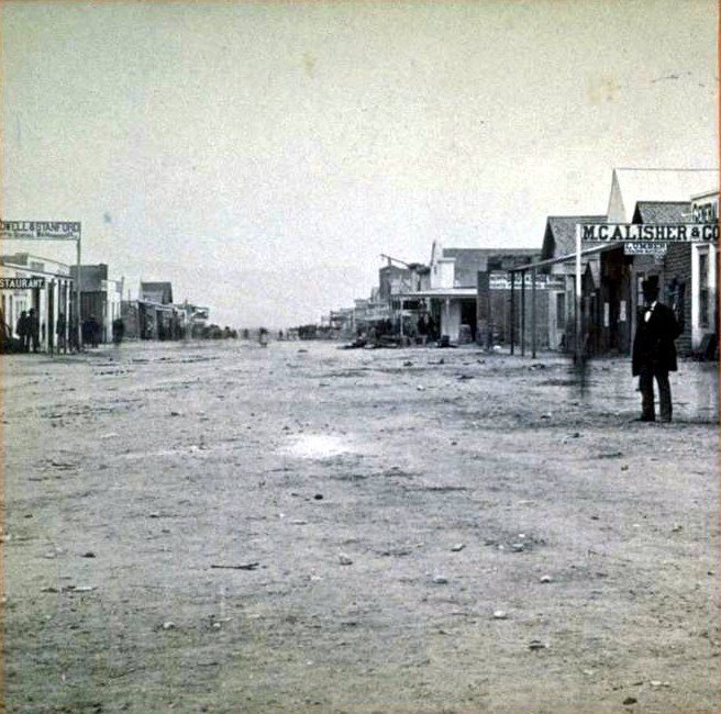 A black and white photograph of a dusty, unpaved main street in Tombstone. Wooden buildings line both sides of the wide dirt road, including storefronts with signs visible such as 'RESTAURANT' and 'McALLISTER & CO'. A lone figure in dark clothing stands on the right side of the street. The scene captures the stark, frontier atmosphere of an early settlement, with simple wooden structures and an empty, expansive street stretching into the distance.